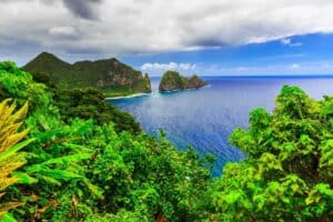 Et fantastisk kystlandskab i Samoa byder på frodig grøn vegetation i forgrunden, krystalklart blåt hav og klippeformationer i det fjerne under en delvist overskyet himmel. Den pulserende scene viser naturens skønhed og tropiske sindsro.