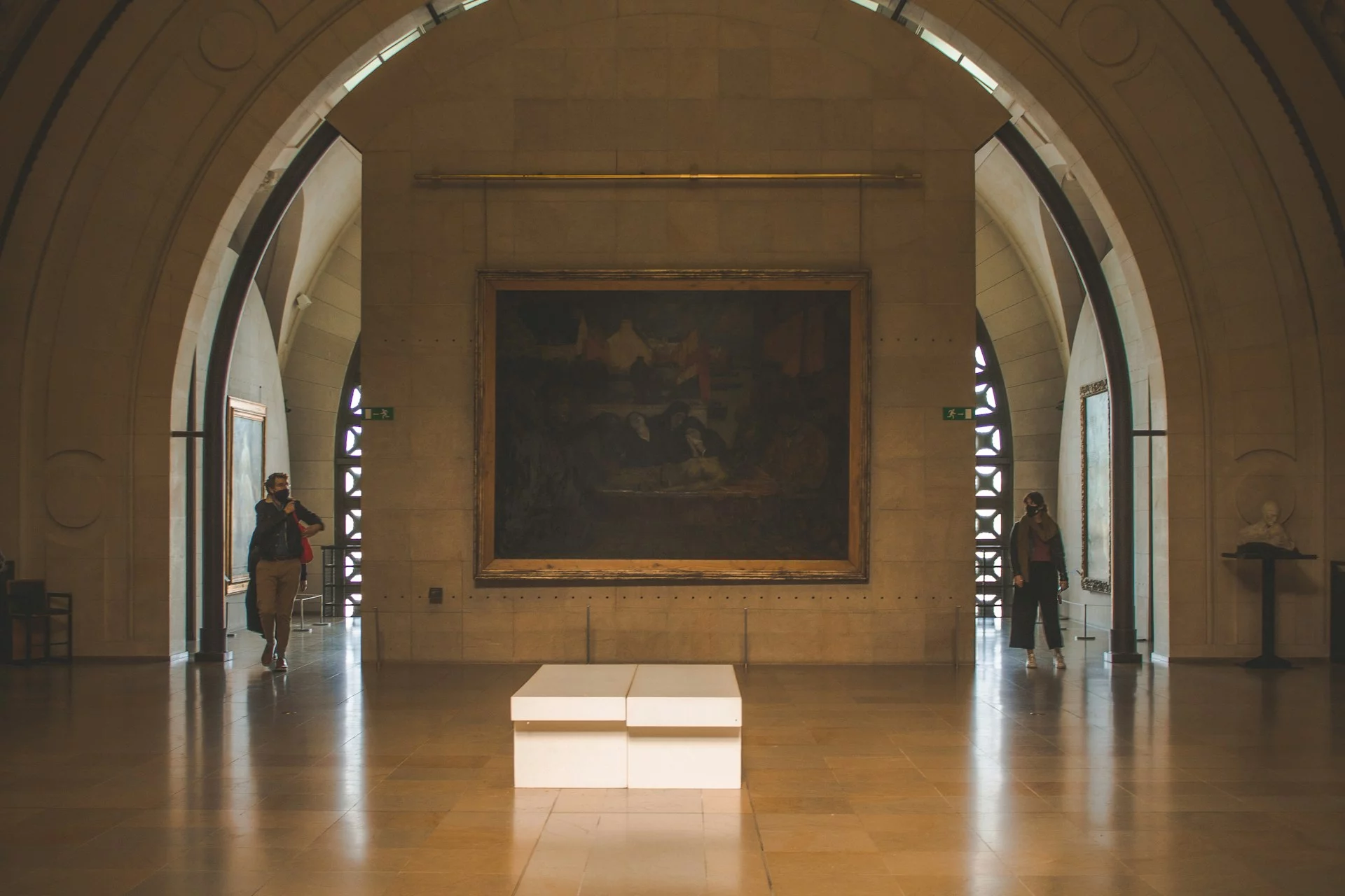 Et stort maleri er vist på en veloplyst væg i et kunstgalleri, der minder om Musée d'Orsay, med buede lofter. To besøgende iført ansigtsmasker står på hver side af kunstværket. Gulvet er lavet af glatte, lyse klinker, og en hvid bænk er placeret foran maleriet.