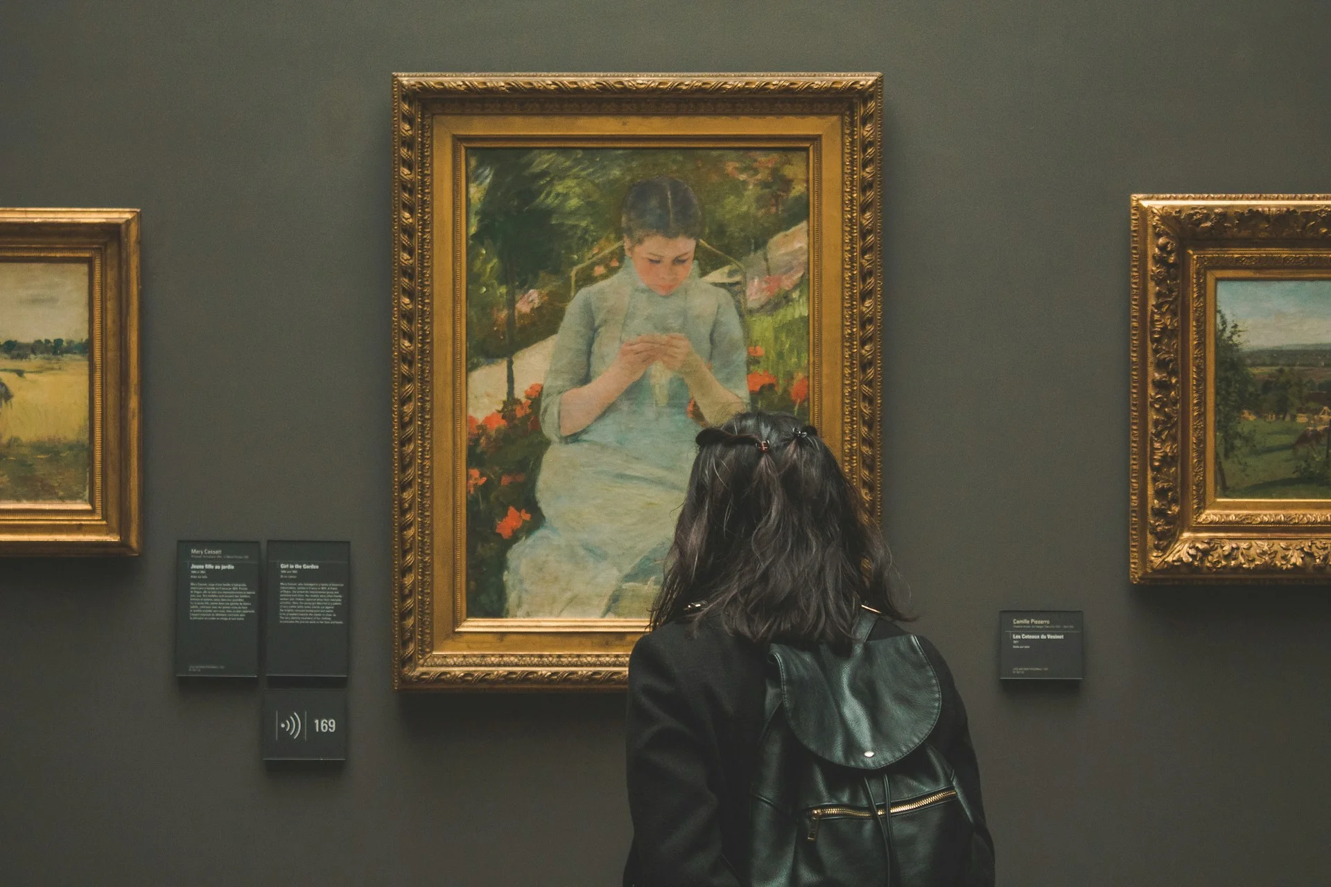 En person med langt, mørkt hår og en sort rygsæk står foran et indrammet maleri i et kunstgalleri, der minder om Musée d'Orsay. Maleriet forestiller en kvinde i blå kjole, der intenst arbejder på en opgave, eventuelt strikker. Andre malerier er synlige på de tilstødende vægge.