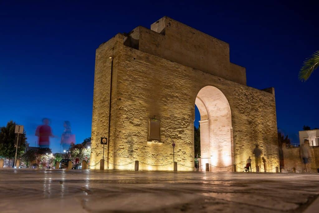Byporten Porta Napoli i Lecce oplyst i den blaa time med mennesker der gaar forbi